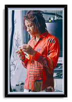 Trippie Redd framed wall art print portrays the rapper with dreadlocks in a red illustrated shirt, looking down as he holds a small object.