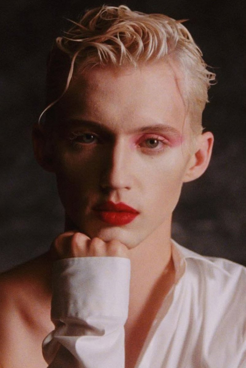 Troye Sivan poster portrait with tousled blond hair, pink eye makeup and red lipstick, resting his chin on a hand.