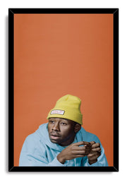 Tyler, The Creator framed wall art print presents his seated portrait in a yellow GOLF beanie and blue hoodie on orange.