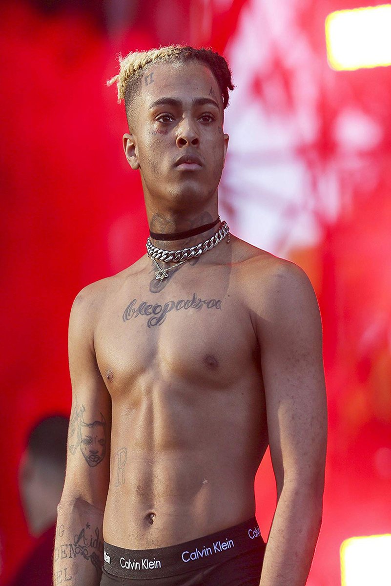 XXXTentacion poster portrait of the shirtless rapper with tattoos and layered chains against a vivid red stage backdrop.